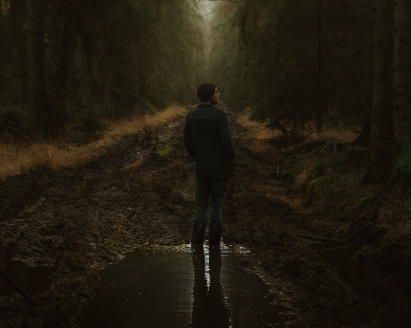 A man stands on a muddy forest path in a dark and atmospheric forest