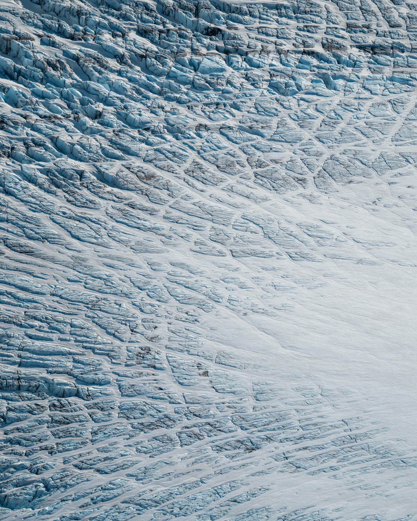 Aerial view of textured glacier ice in Alaska with intersecting cracks and subtle blue tones