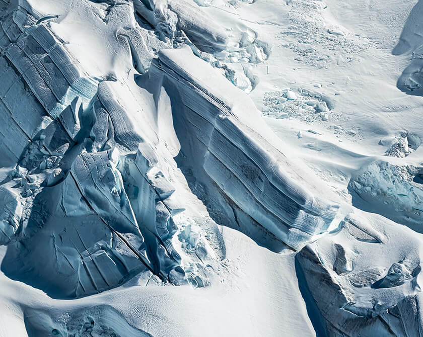 Aerial view of textured glacier ice with intersecting cracks and subtle blue tones.