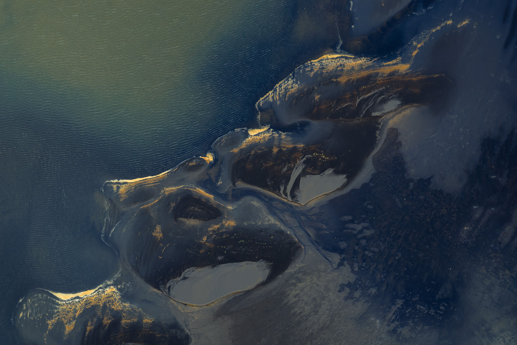 Aerial view of Icelandic lagoon with golden foam patterns on dark sand and deep blue water
