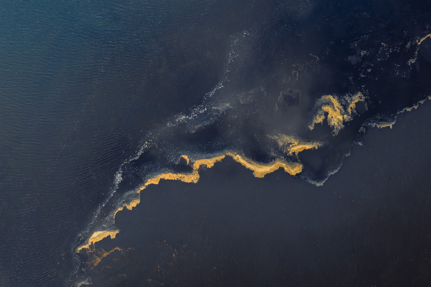 Aerial view of lagoon surface in Iceland with gold-tinted foam drifting across dark water
