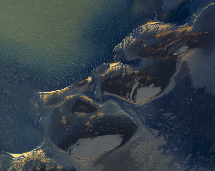 Aerial view of Icelandic lagoon where golden foam traces curve across deep blue tidal water